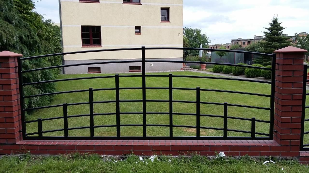 Czarny, metalowy płot na niskiej, ceglanej podmurówce z ceglanymi słupkami, z widokiem na trawnik i budynek mieszkalny w tle. Płot o łukowatym kształcie.