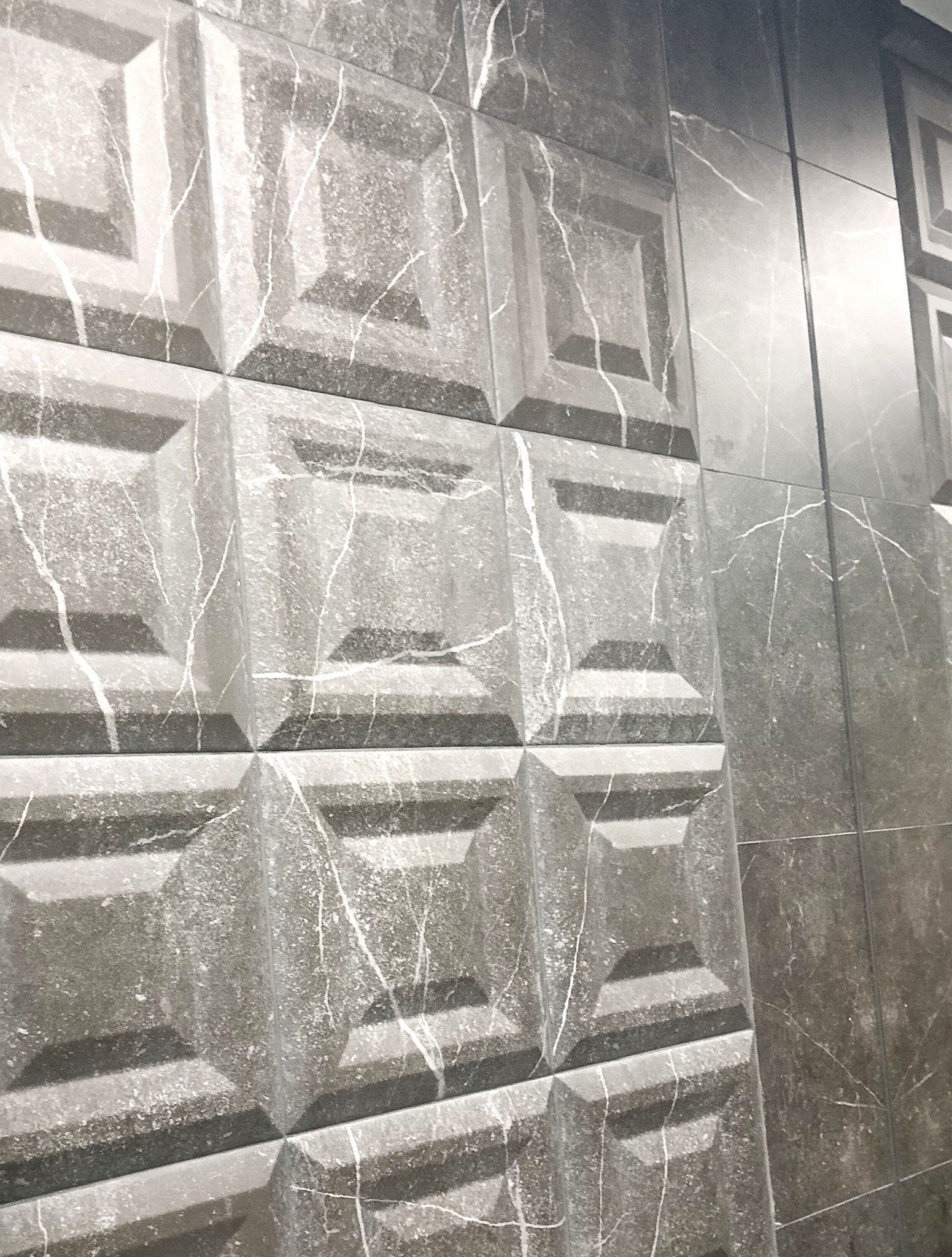 Szara, trójwymiarowa okładzina ścienna z efektem marmuru i wyraźną fakturą. Ułożona w regularny wzór, z widocznymi fugami. Część płytek poza kadrem.