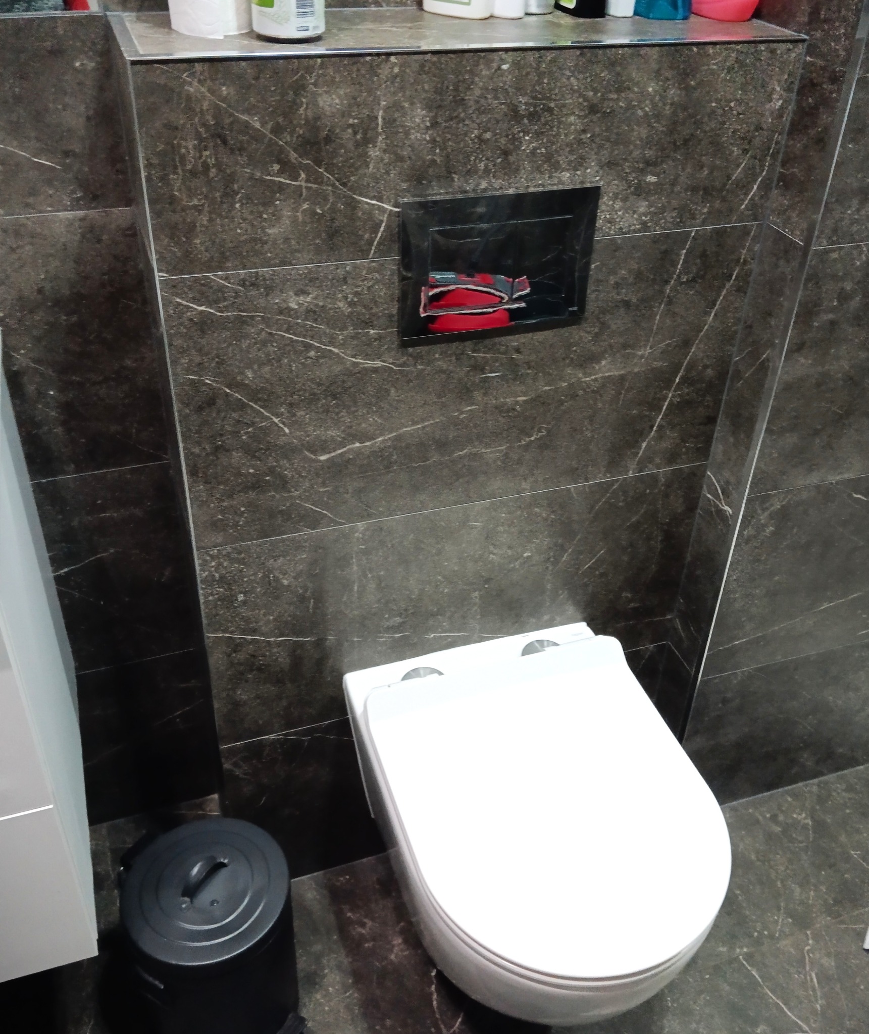 Wykończona łazienka z toaletą podwieszaną i ciemnymi płytkami imitującymi marmur. Widoczny czarny kosz na śmieci i przycisk spłuczki w czarnej ramce.