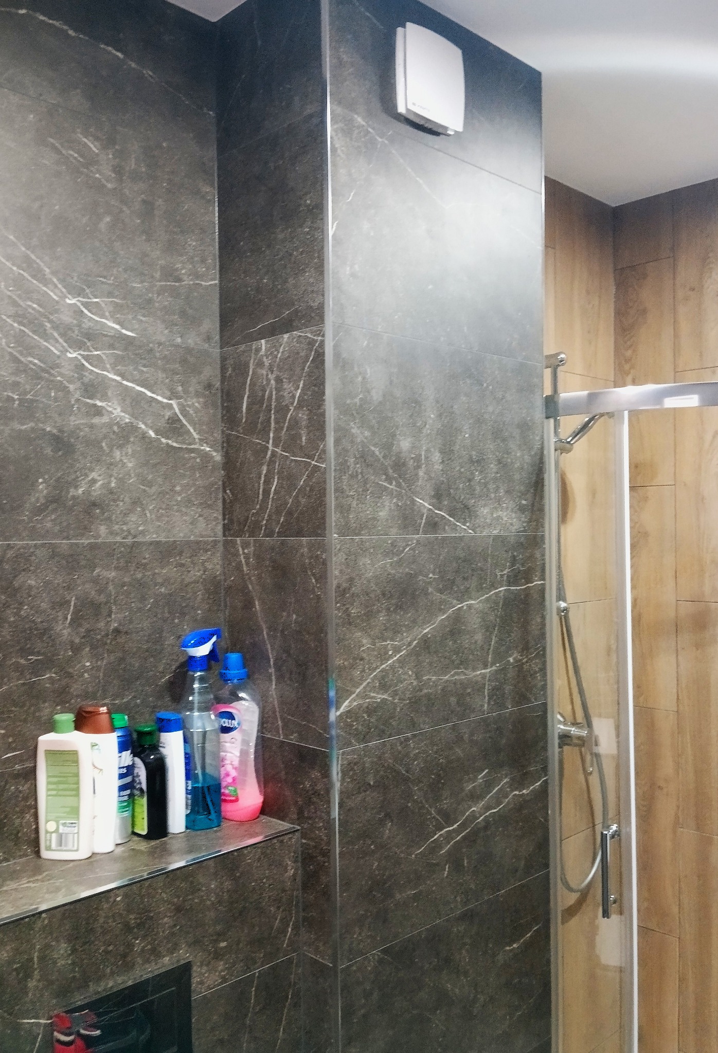 Wykończona łazienka z szarymi płytkami imitującymi marmur, kabiną prysznicową i półką z kosmetykami. Widoczny wentylator na ścianie.