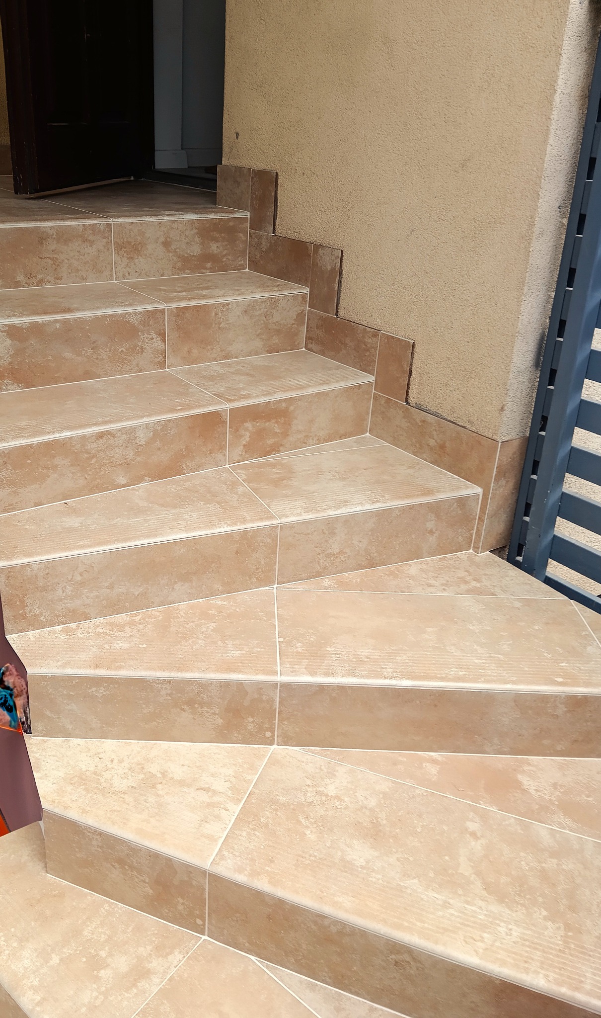 Wykończone schody zewnętrzne z beżowych płytek, z widocznymi fugami i fragmentem ściany o podobnej kolorystyce. Ujęcie z dołu, podkreślające detale wykończenia.