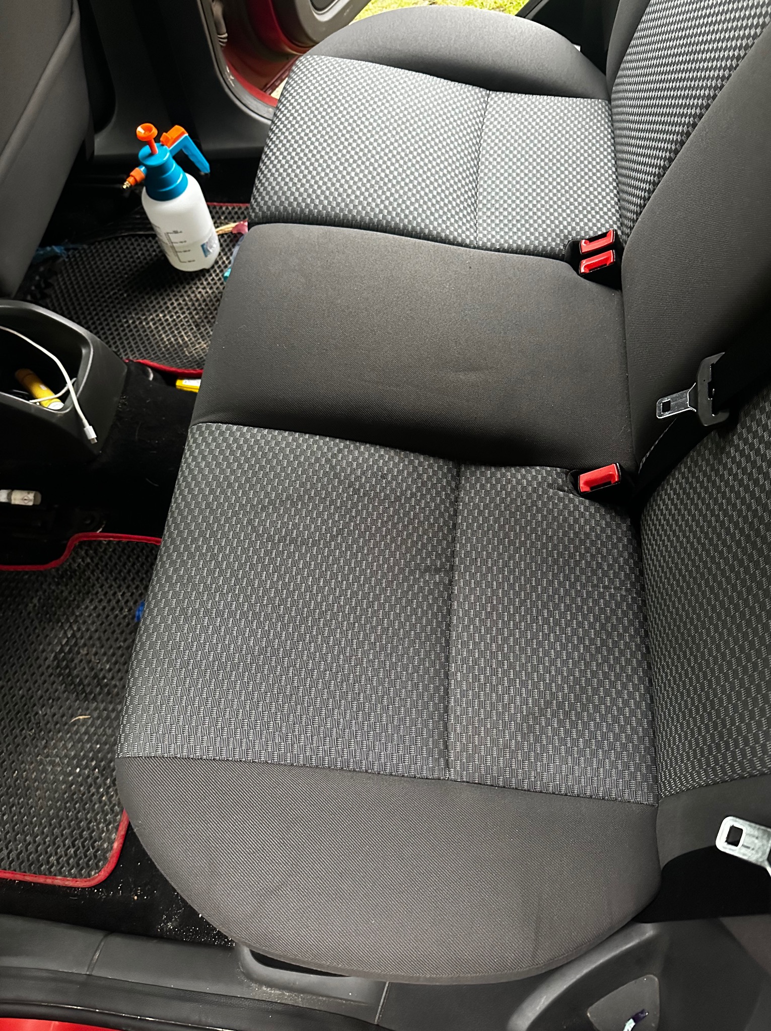 Wnętrze samochodu przed czyszczeniem: tylna kanapa z widocznymi zabrudzeniami i butelką ze środkiem czyszczącym. Dywaniki z czerwonym obramowaniem.