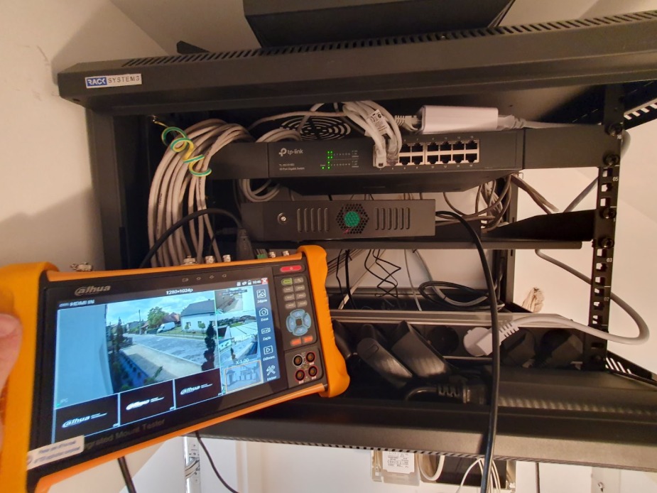 Tester w dłoni wyświetla obraz z kamer monitoringu na tle szafy rack z urządzeniami sieciowymi. Widoczne kable, switch i inne elementy instalacji monitoringu.