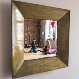 kaskal - Katarzyna Lichnowska - Dwie kobiety siedzą na podłodze w jasnym pokoju z ceglaną ścianą, widoczne w lustrze o złotej ramie. Jedna gestykuluje, druga słucha. Naturalne światło z okna.