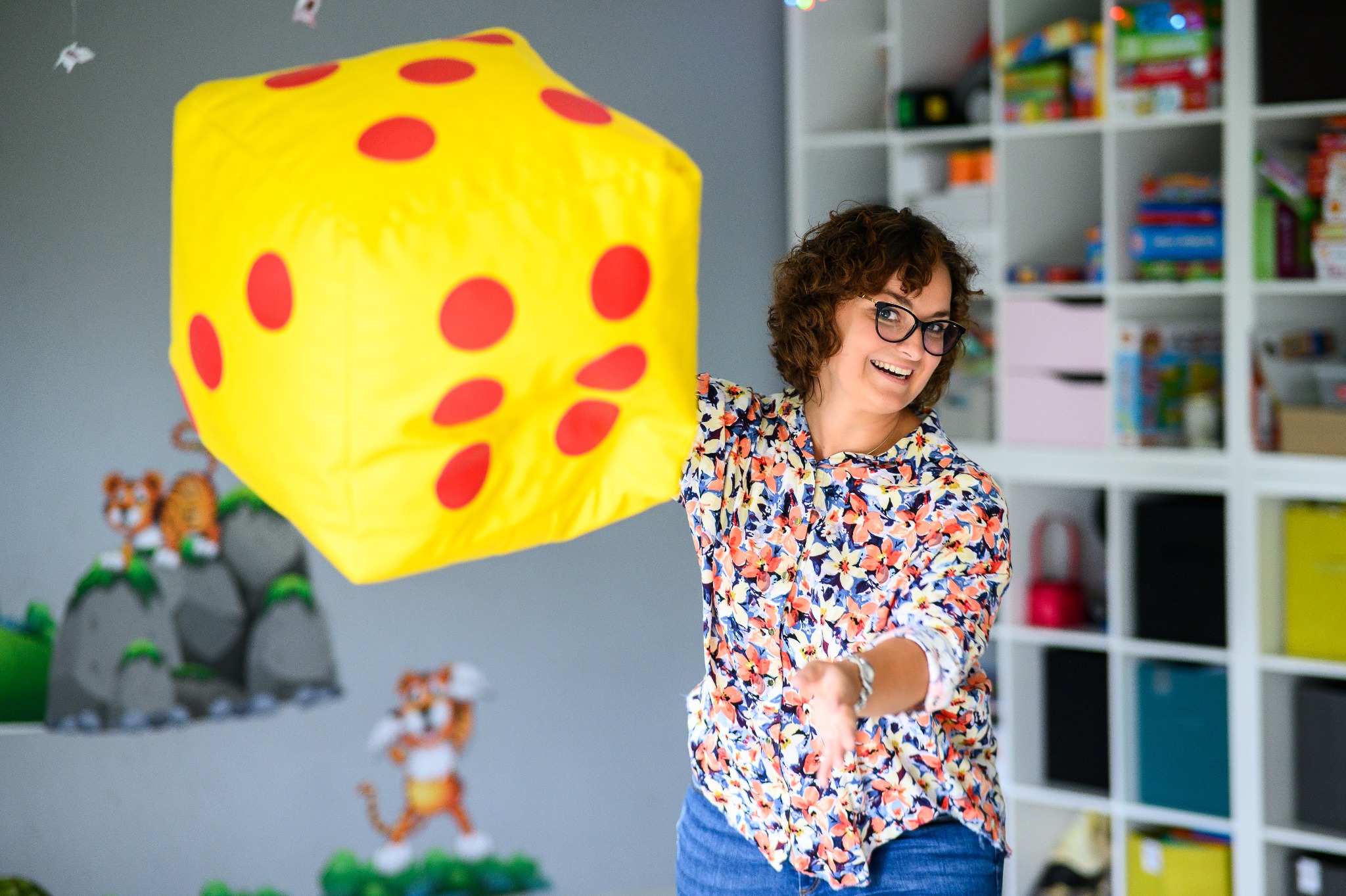 Uśmiechnięta kobieta w okularach trzyma dużą, żółtą kostkę z czerwonymi kropkami na tle pokoju dziecięcego z półkami pełnymi zabawek i rysunkami na ścianie.
