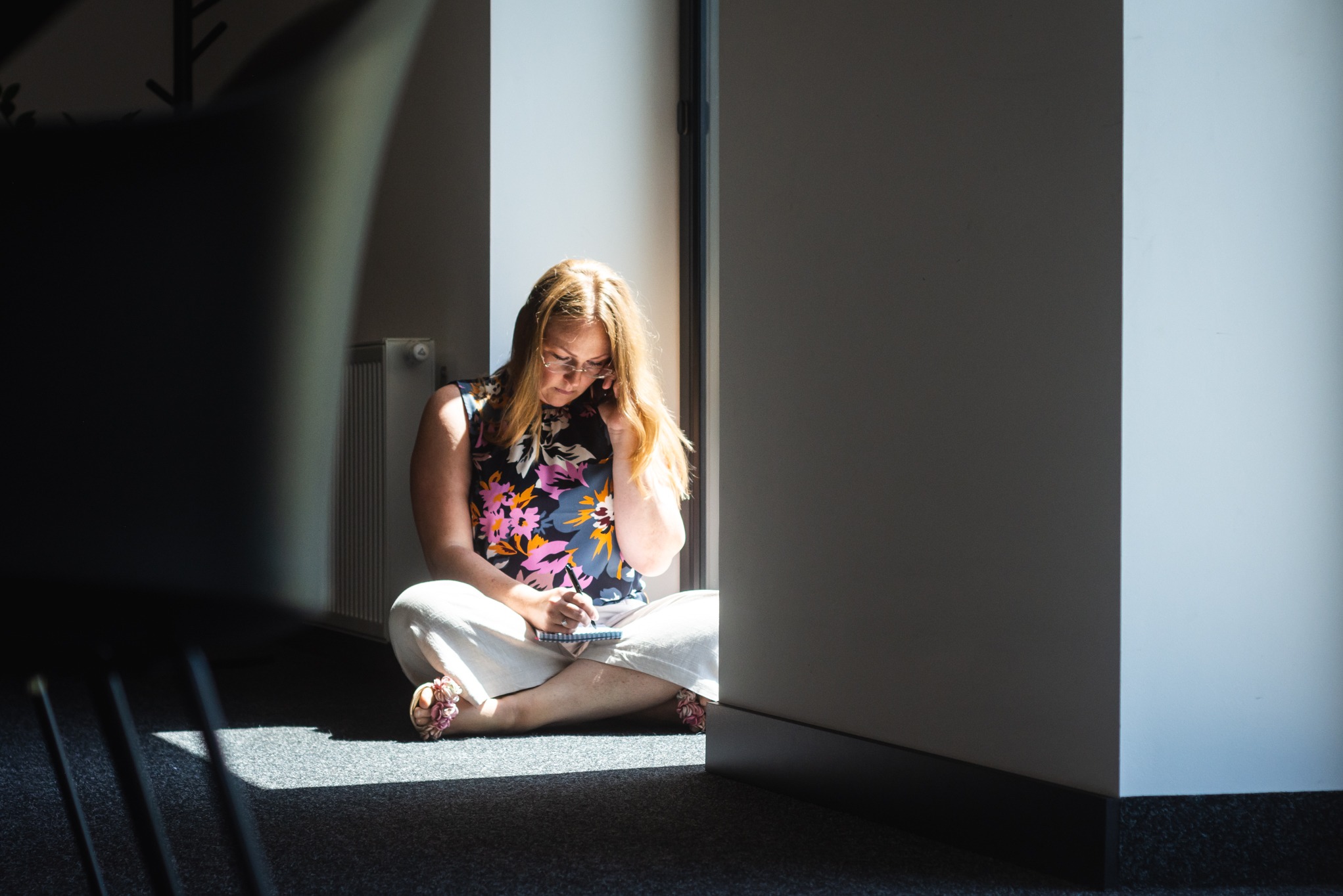 Kobieta w kwiecistej bluzce siedzi na podłodze w jasnym świetle, pisząc w notatniku. W tle widoczny fragment ściany i grzejnik.