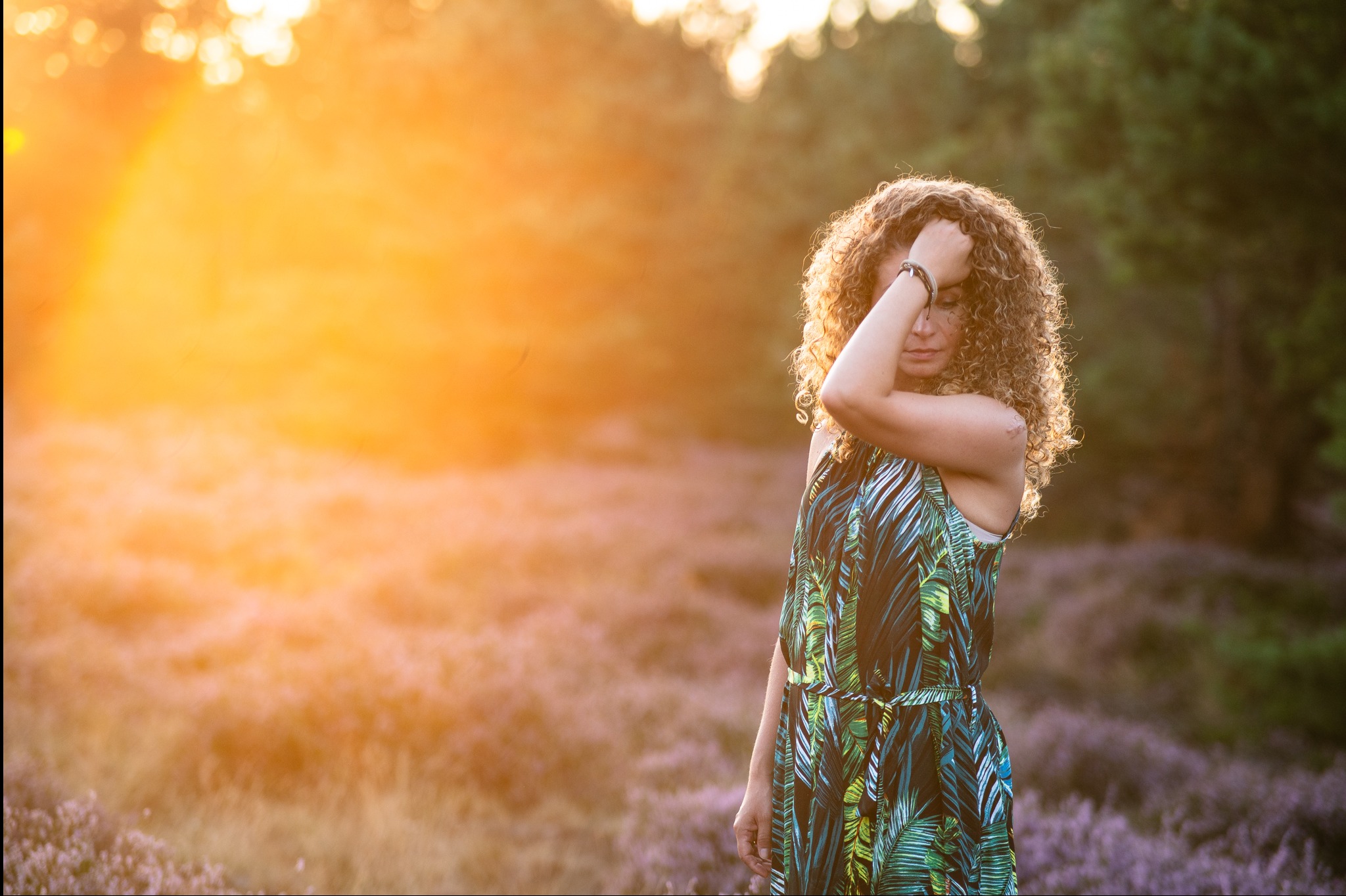 Kobieta z kręconymi włosami w sukience w tropikalne liście, stojąca na tle kwitnącego wrzosowiska w promieniach zachodzącego słońca. Delikatne światło podkreśla naturalność sceny.