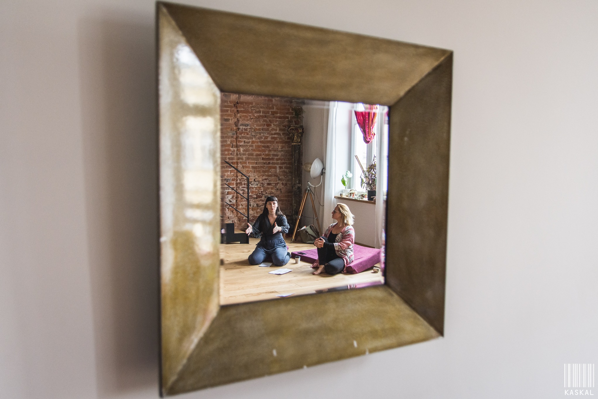 Dwie kobiety siedzą na podłodze w jasnym pokoju z ceglaną ścianą, widoczne w lustrze o złotej ramie. Jedna gestykuluje, druga słucha. Naturalne światło z okna.