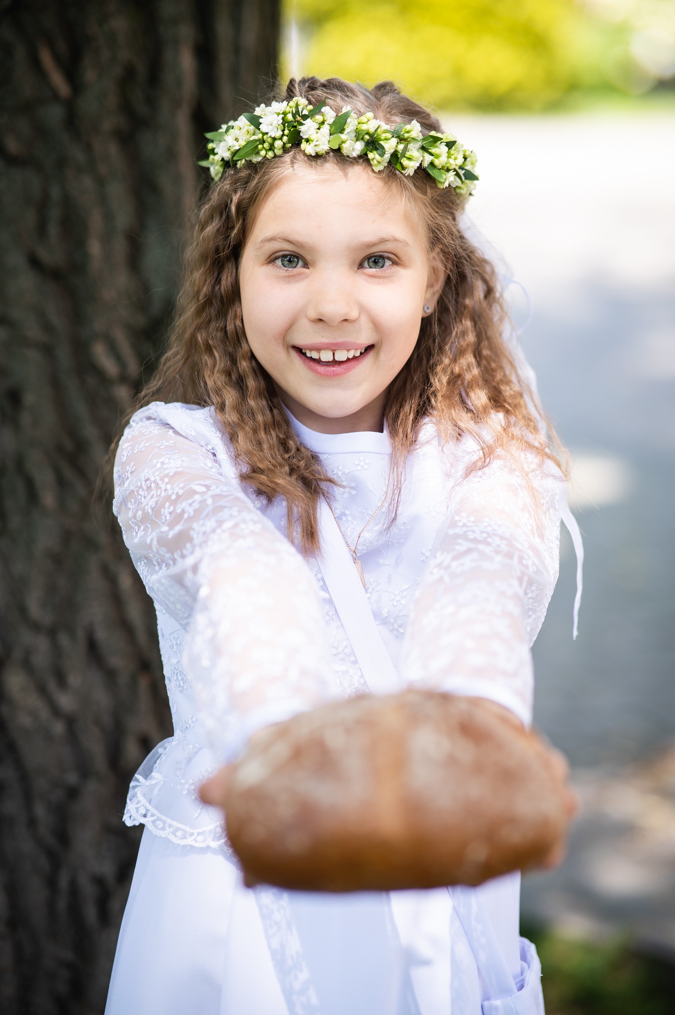 Uśmiechnięta dziewczynka w białej sukience komunijnej i wianku, wyciąga ręce z chlebem w stronę obiektywu na tle drzewa i rozmytego tła.