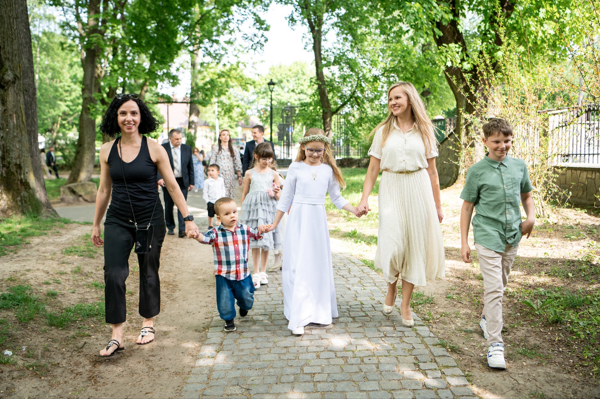 Uroczystość Pierwszej Komunii Świętej: Grupa dzieci i dorosłych idzie ścieżką w parku, dziewczynka w białej sukience komunijnej na pierwszym planie.