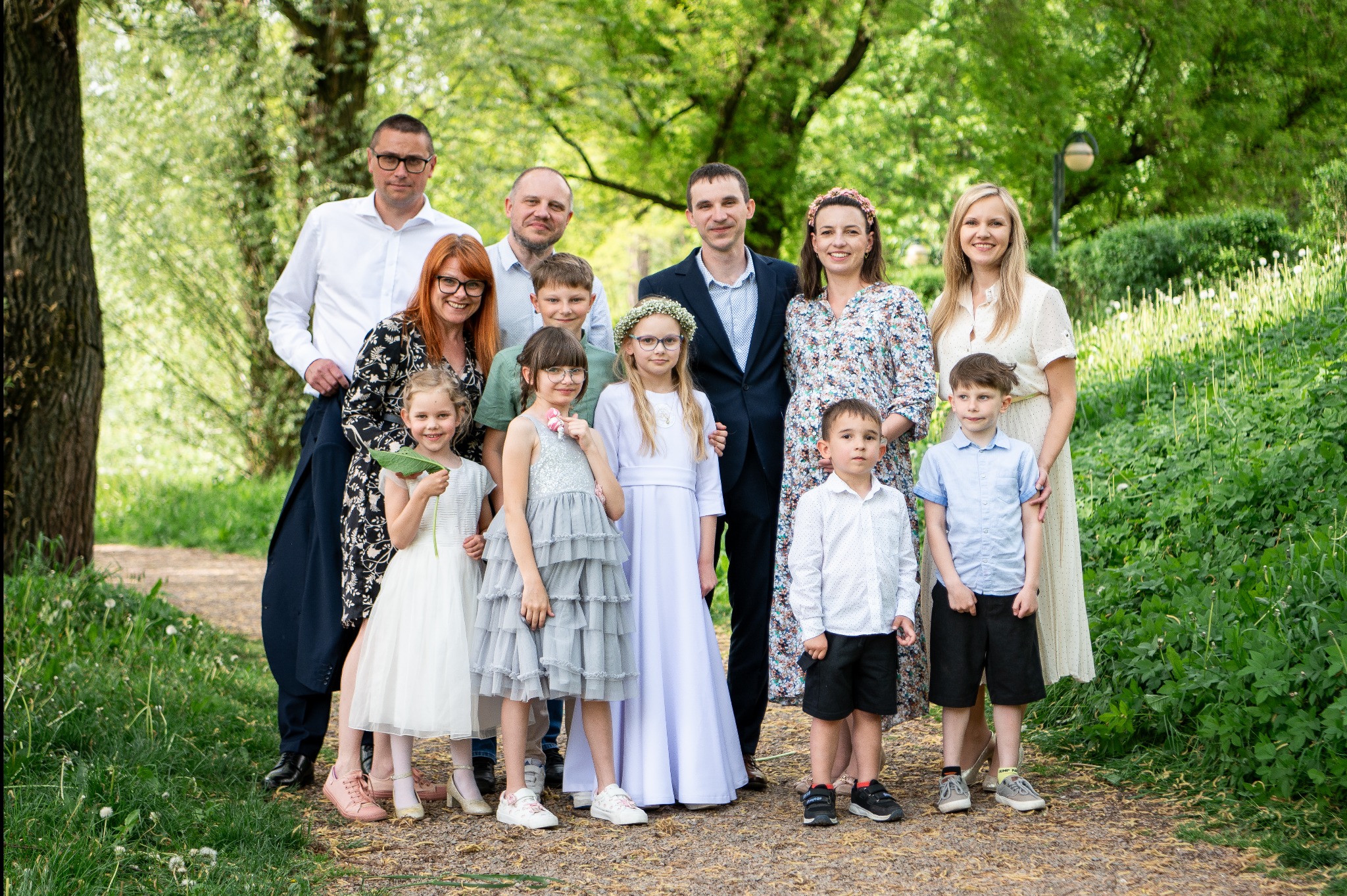 Duża, uśmiechnięta rodzina pozuje do zdjęcia w plenerze. Dzieci ubrane w eleganckie stroje, w tle drzewa i zieleń. Fotografia rodzinna w naturalnym otoczeniu.