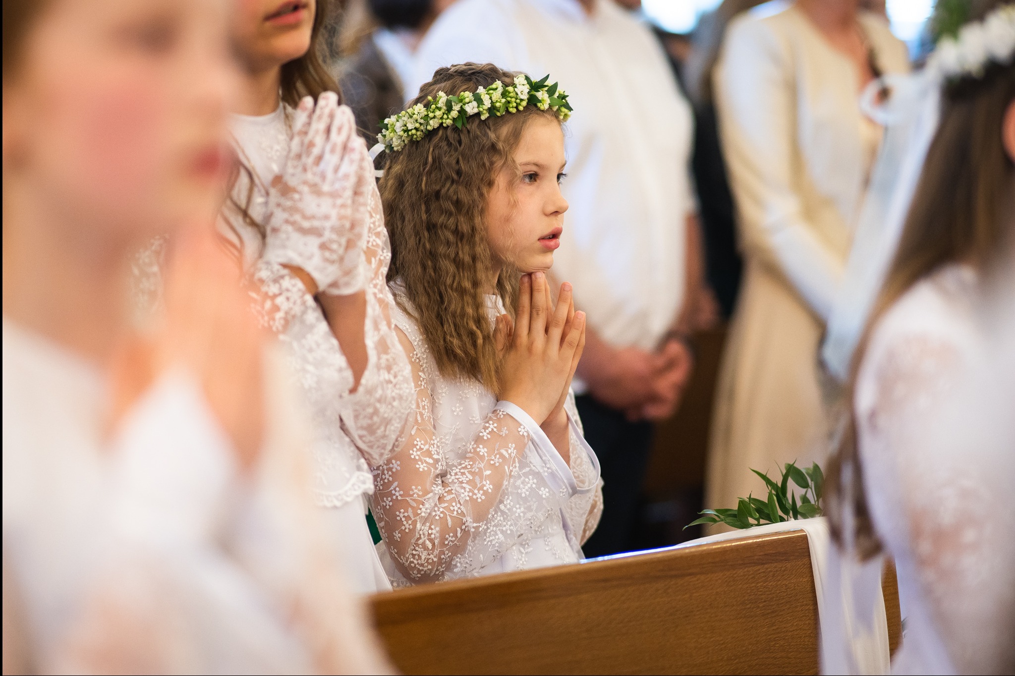 Dziewczynka w białej sukience i wianku na głowie, z złożonymi rękami, uczestniczy w uroczystości religijnej. W tle widoczni inni uczestnicy.