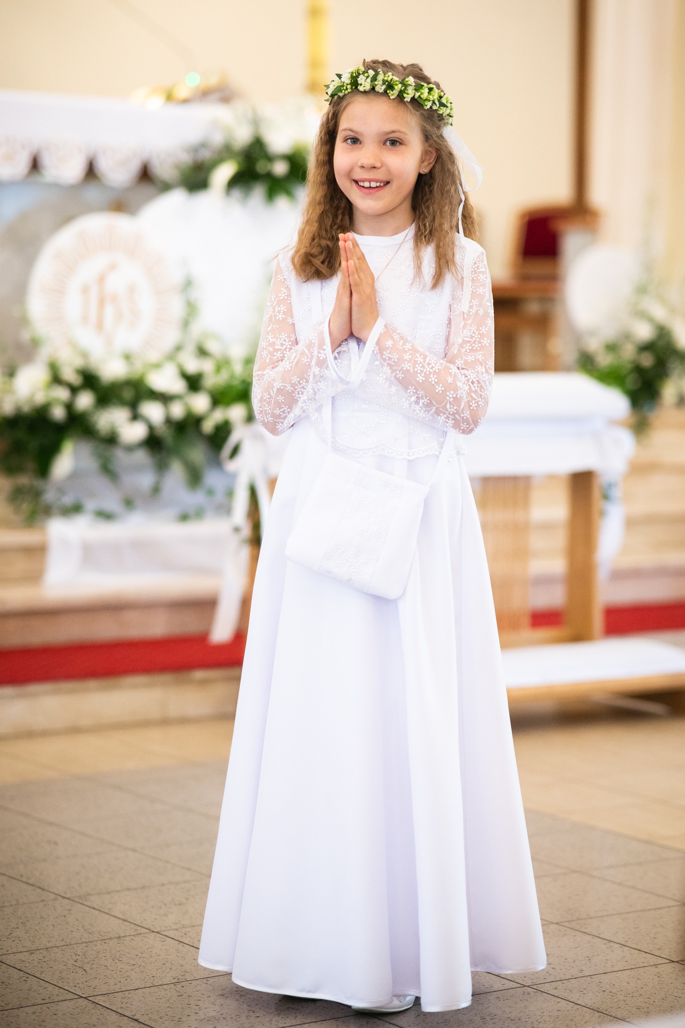 Uśmiechnięta dziewczynka w białej sukience komunijnej, z wiankiem na głowie, stoi w kościele z złożonymi rękami. Delikatne światło, jasne tło.