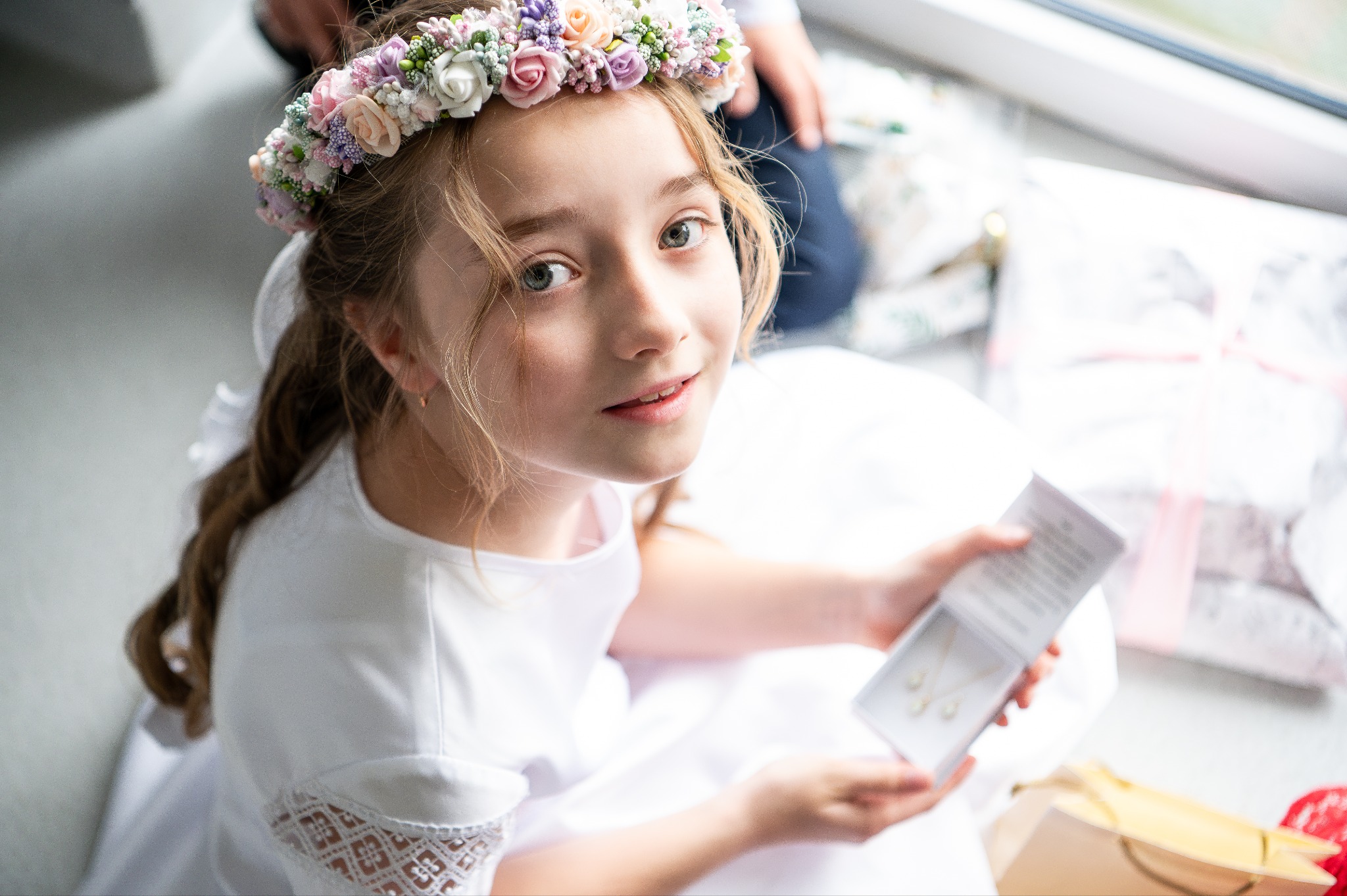 Dziewczynka w białej sukience i wianku z kwiatów, siedząca przy oknie, trzymająca pudełko z biżuterią. Uśmiechnięta, patrzy w górę. Jasne, naturalne światło.