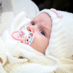 Studio4U - Noworodek z niebieskimi oczami, w białym, dzierganym czepku z różową kokardką, leży otulony w miękkim kocyku, z smoczkiem w ustach. Delikatne światło podkreśla detale.