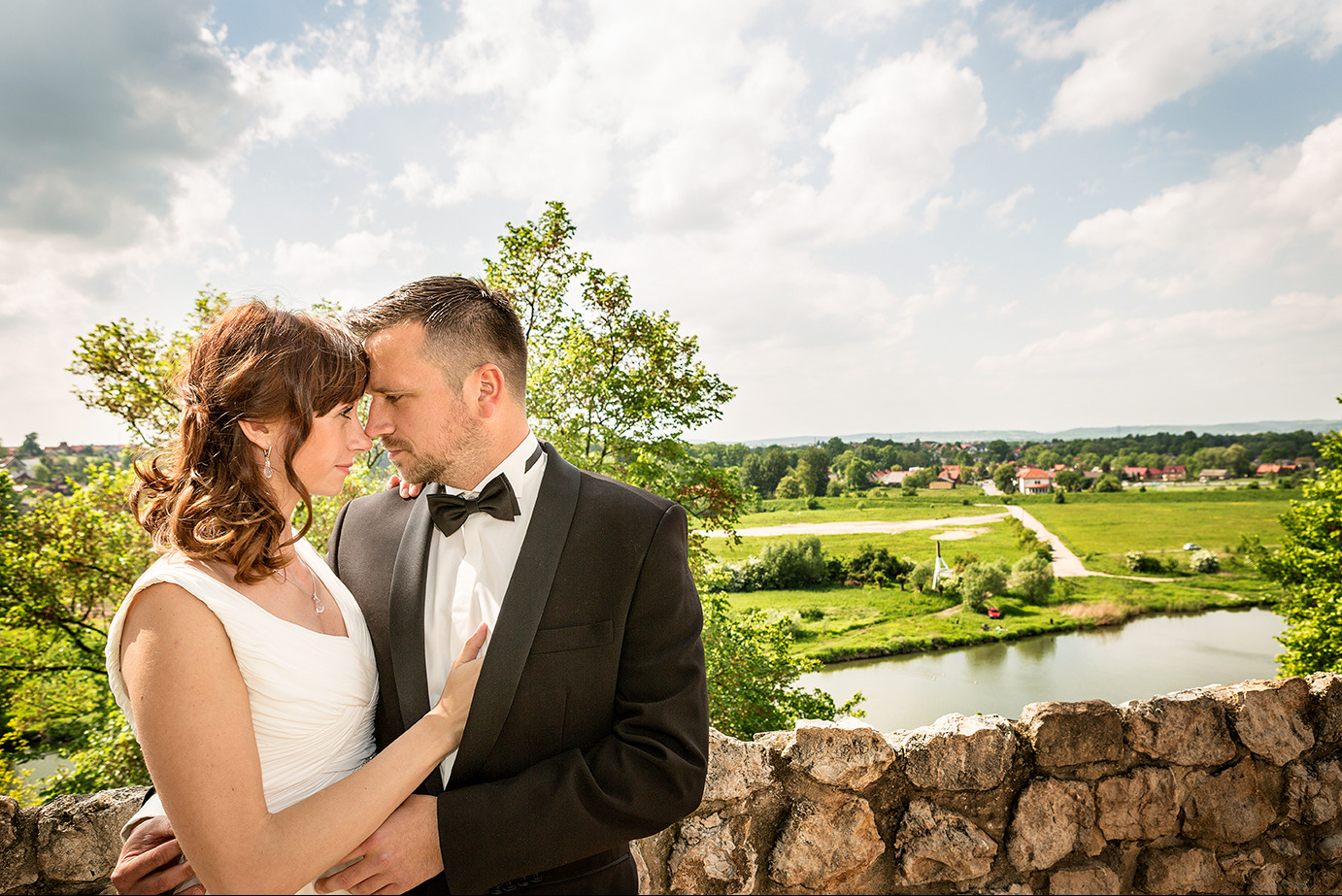 Para młoda w objęciach na tle malowniczego krajobrazu z jeziorem i zielenią. Pan młody w smokingu, panna młoda w białej sukni. Romantyczna atmosfera.