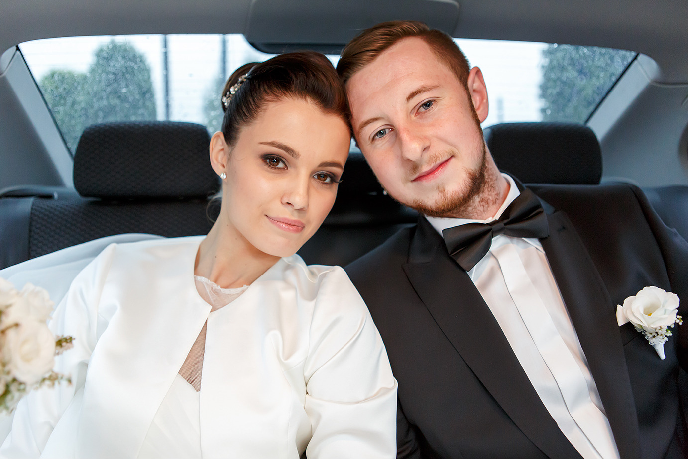 Uśmiechnięta para młoda w samochodzie: panna młoda w białej sukni i żakiecie, pan młody w smokingu z muchą. Widoczne kwiaty i tapicerka auta.