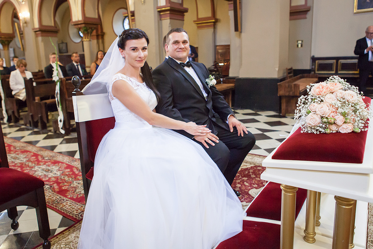 Uśmiechnięta para młoda siedzi w kościele, trzymając się za ręce. Bukiet róż na stole, czarno-biała podłoga i goście w tle. Ujęcie z boku, naturalne światło.