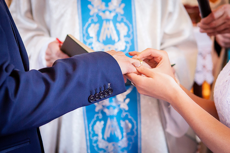 Moment zakładania obrączki podczas ceremonii ślubnej. Widoczny fragment sukni panny młodej, garnitur pana młodego i strój księdza.