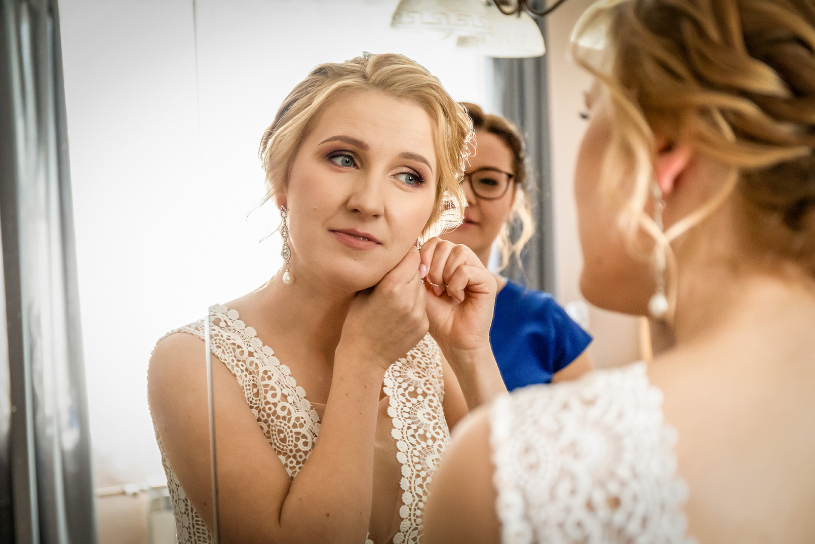 Panna młoda w koronkowej sukni poprawia kolczyk przed lustrem. W tle wizażystka w niebieskiej bluzce. Delikatny makijaż i upięcie włosów.