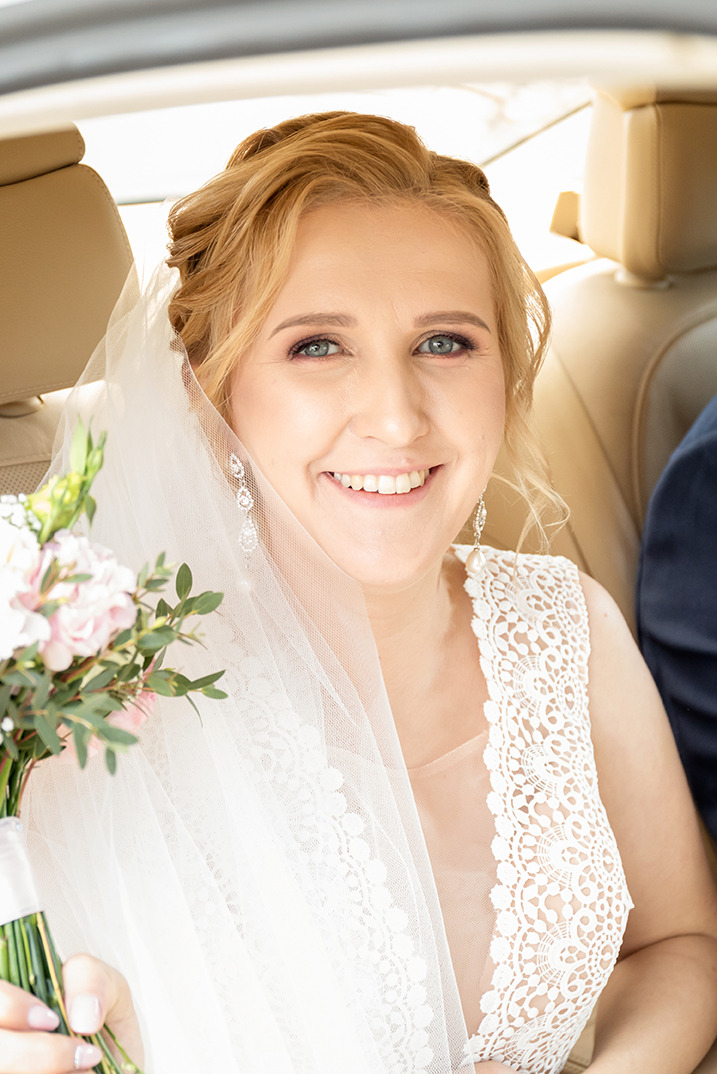 Uśmiechnięta panna młoda w koronkowej sukni i welonie, siedząca w samochodzie, trzyma bukiet z różowymi kwiatami. Widoczne kolczyki i fragment marynarki pana młodego.
