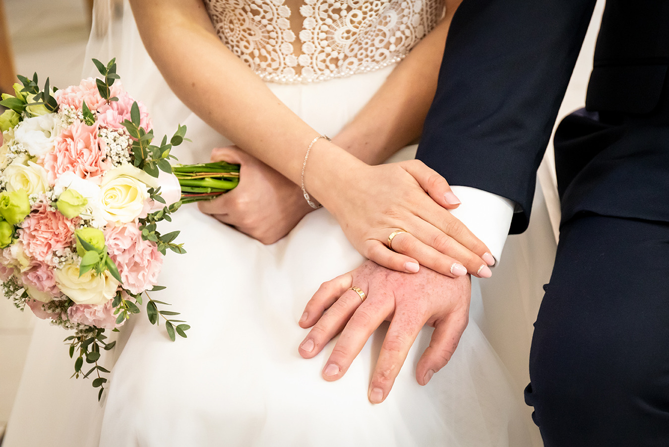 Dłonie pary młodej z obrączkami na tle sukni ślubnej i bukietu z różowych i białych kwiatów. Delikatny makijaż panny młodej i elegancki garnitur pana młodego.