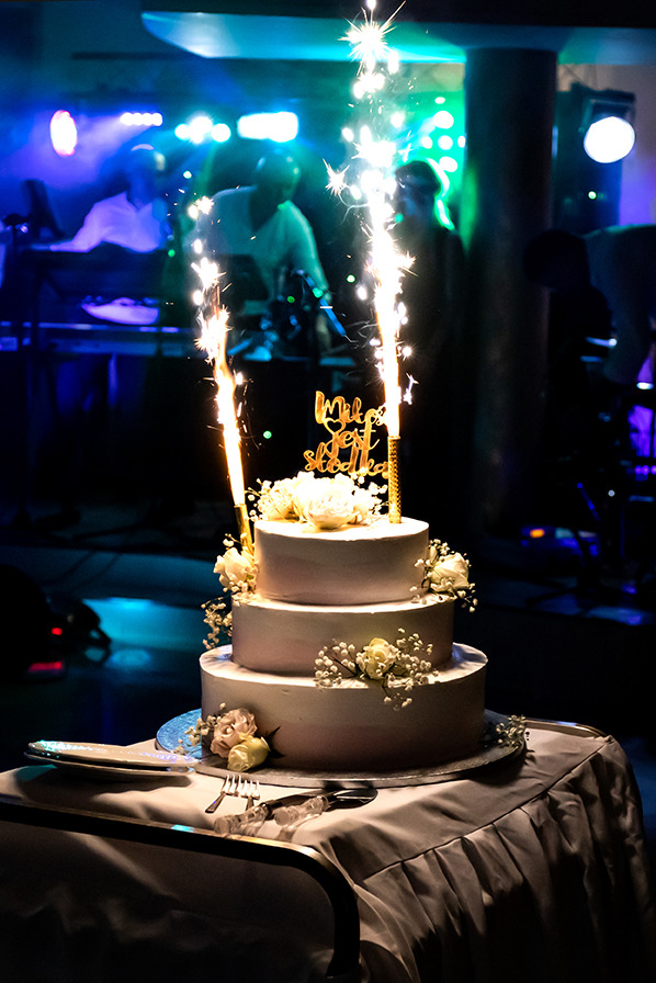 Trzypiętrowy tort weselny z białymi różami i fontannami iskier na stole z białym obrusem, w tle zespół muzyczny i oświetlenie sceniczne.