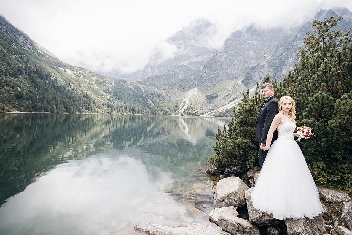 Para młoda w eleganckich strojach pozuje na tle górskiego jeziora z mglistymi szczytami. Ujęcie z dołu podkreśla majestat krajobrazu i uroczysty charakter chwili.