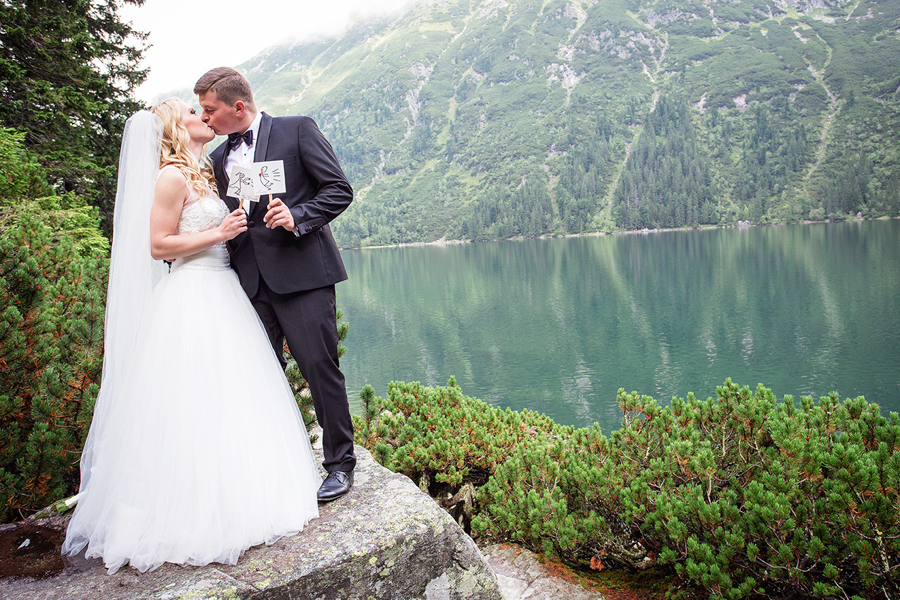 Pocałunek nowożeńców na tle górskiego jeziora. Pan młody trzyma kartkę z rysunkiem pary. Romantyczna sesja ślubna w plenerze, naturalne światło.