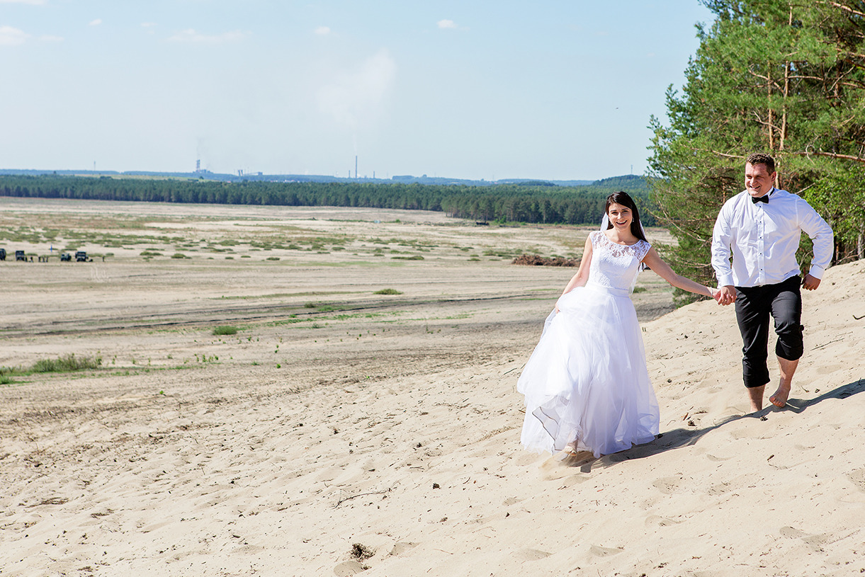 Para młoda, boso, trzymając się za ręce, biegnie po piaszczystej wydmie. W tle las i zabudowania przemysłowe. Jasne, słoneczne światło.
