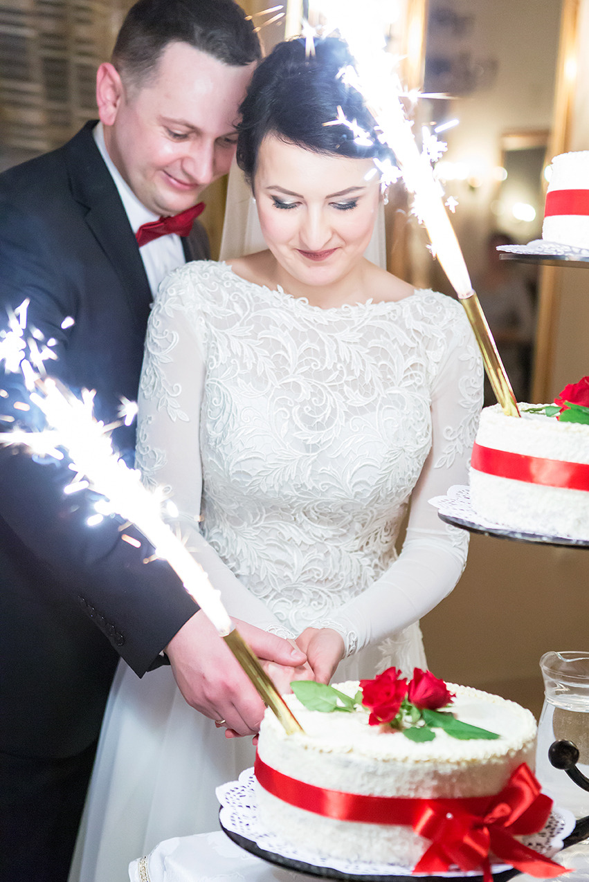 Uśmiechnięta para młoda kroi tort weselny, ozdobiony czerwoną wstążką i różami, z płonącymi zimnymi ogniami. Eleganckie wnętrze sali.