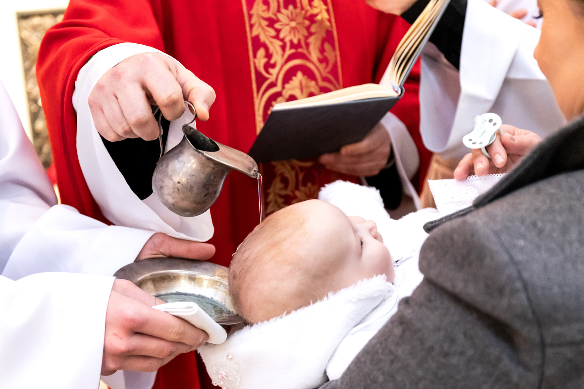 Chrzest dziecka: ksiądz polewa wodą główkę niemowlęcia, trzymanego przez rodziców. W tle otwarta księga i czerwona szata liturgiczna.