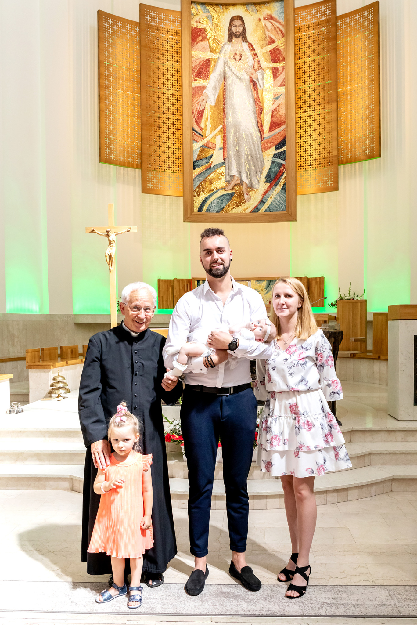 Uroczystość chrztu w kościele: rodzina z dzieckiem, ksiądz i dziewczynka pozują na tle ołtarza z obrazem Jezusa Miłosiernego i złotym krzyżem.