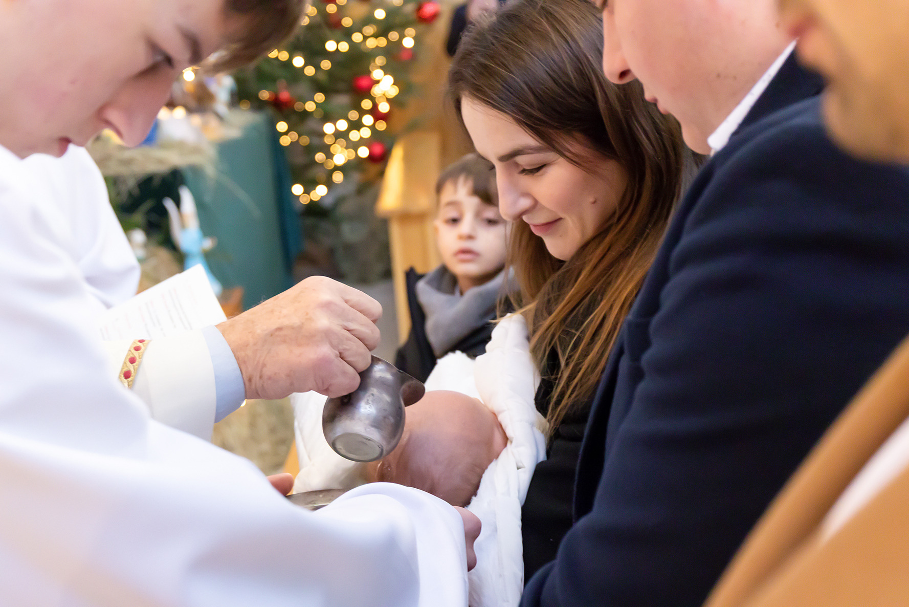 Chrzest dziecka: ksiądz polewa wodą główkę niemowlęcia trzymanego przez matkę, w tle choinka i dziecko obserwujące ceremonię. Ujęcie z bliska, ciepłe światło.