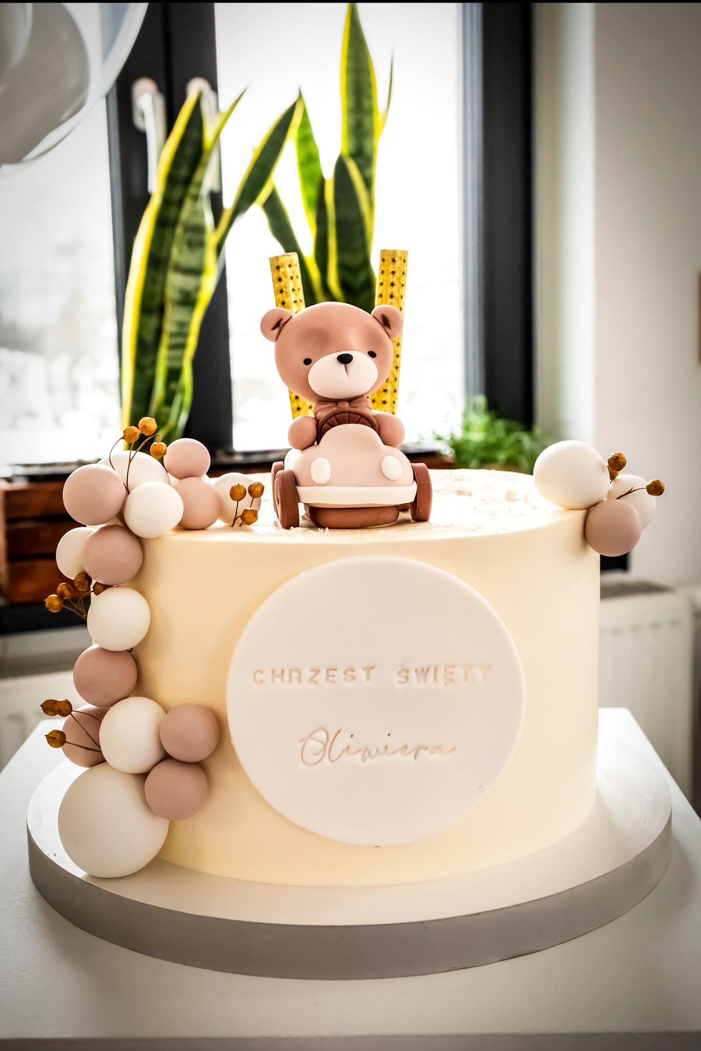 Tort na chrzest z misiem w samochodzie i napisem 'Chrzest Święty Oliwiera', dekorowany kulkami i roślinnymi akcentami, na tle okna z zieloną rośliną.