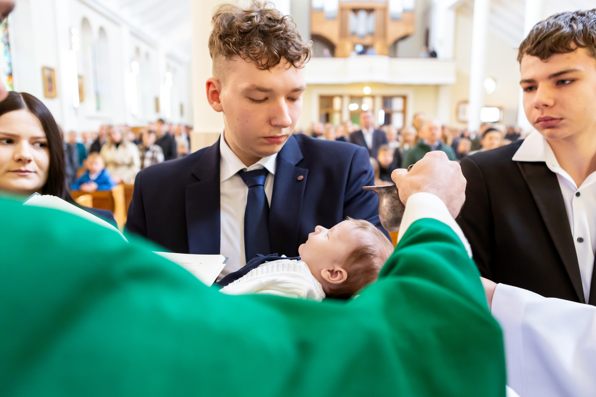Chrzest dziecka w kościele: ksiądz polewa wodą główkę niemowlęcia, w tle rodzina i goście. Uroczystość w jasnym wnętrzu z drewnianymi elementami.