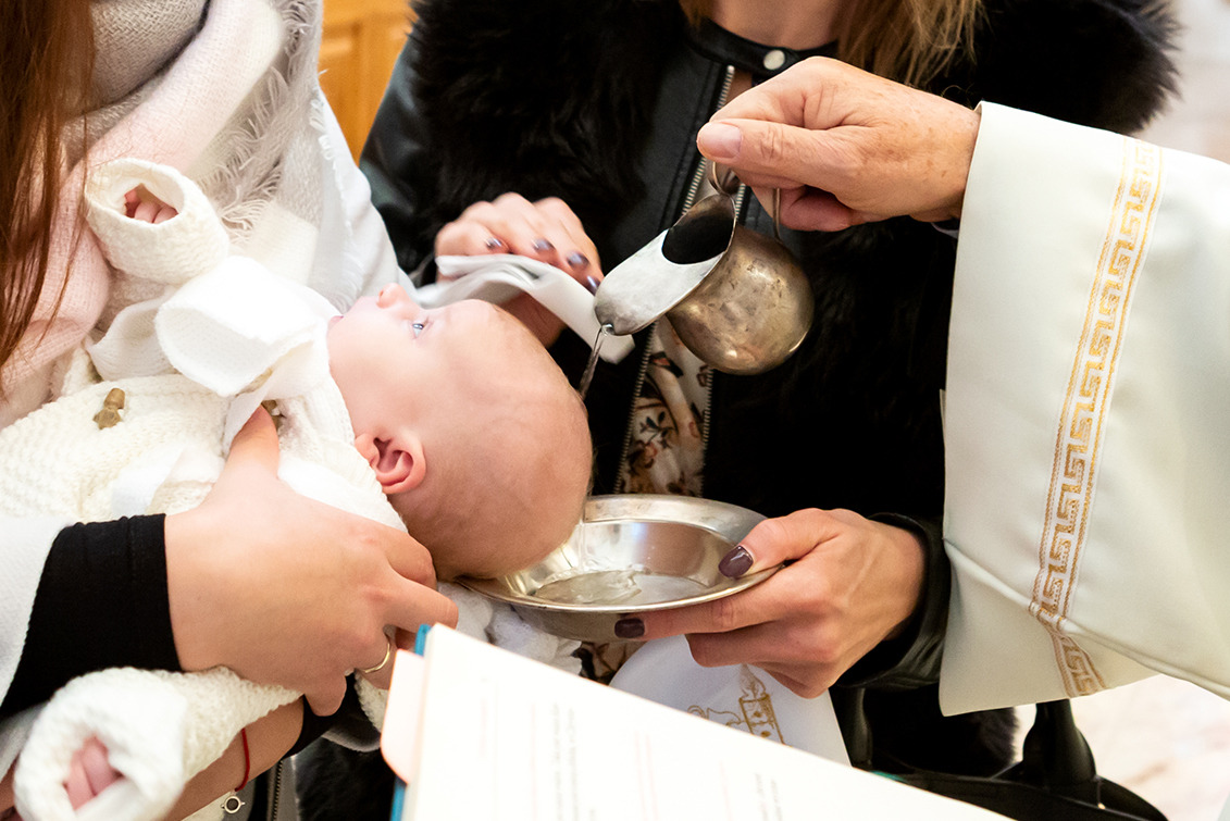 Chrzest dziecka: woda z naczynia wylewana na główkę niemowlęcia trzymanego przez dwie osoby, w tle ksiądz w białej szacie. Ujęcie z bliska, ciepłe światło.