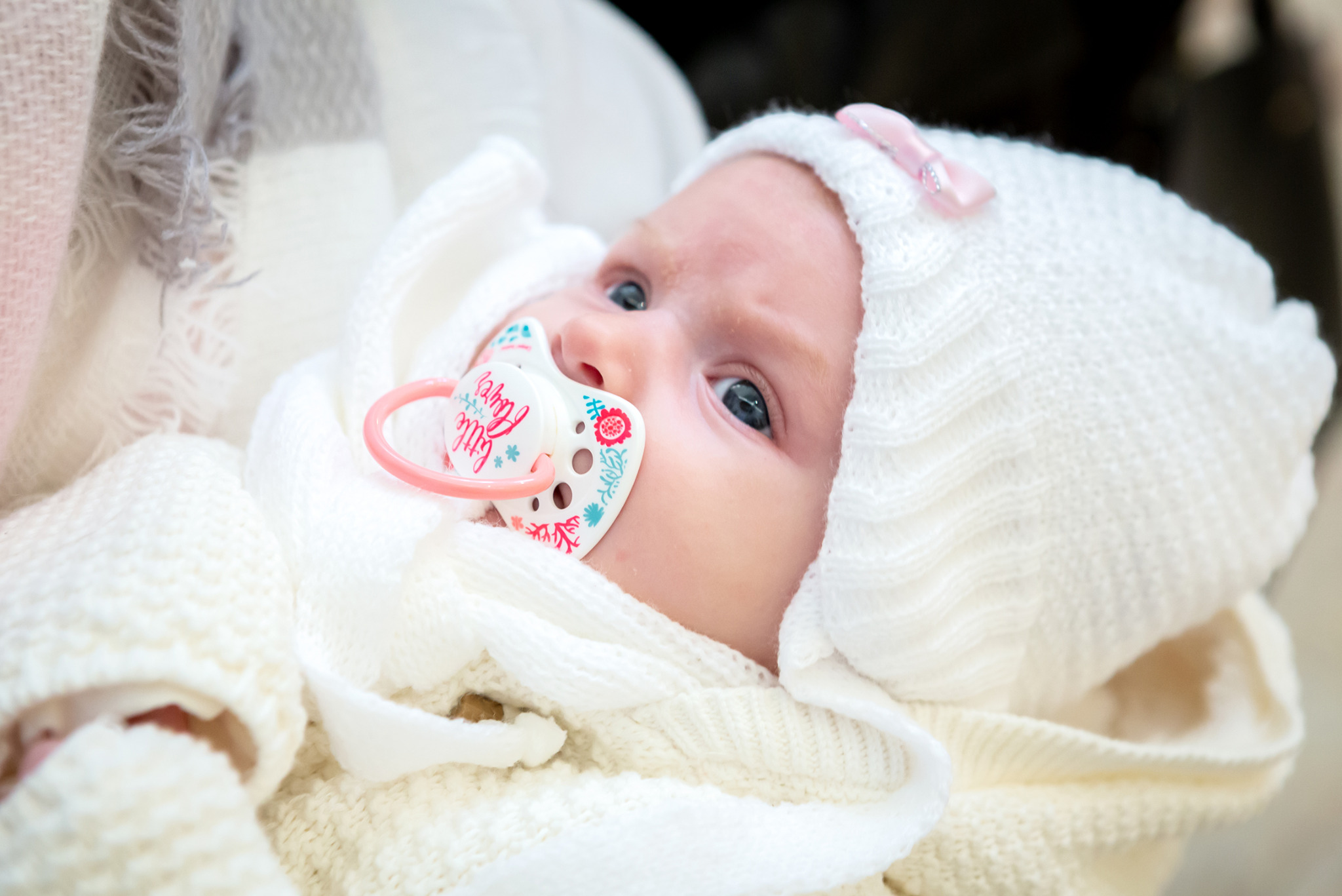 Noworodek z niebieskimi oczami, w białym, dzierganym czepku z różową kokardką, leży otulony w miękkim kocyku, z smoczkiem w ustach. Delikatne światło podkreśla detale.
