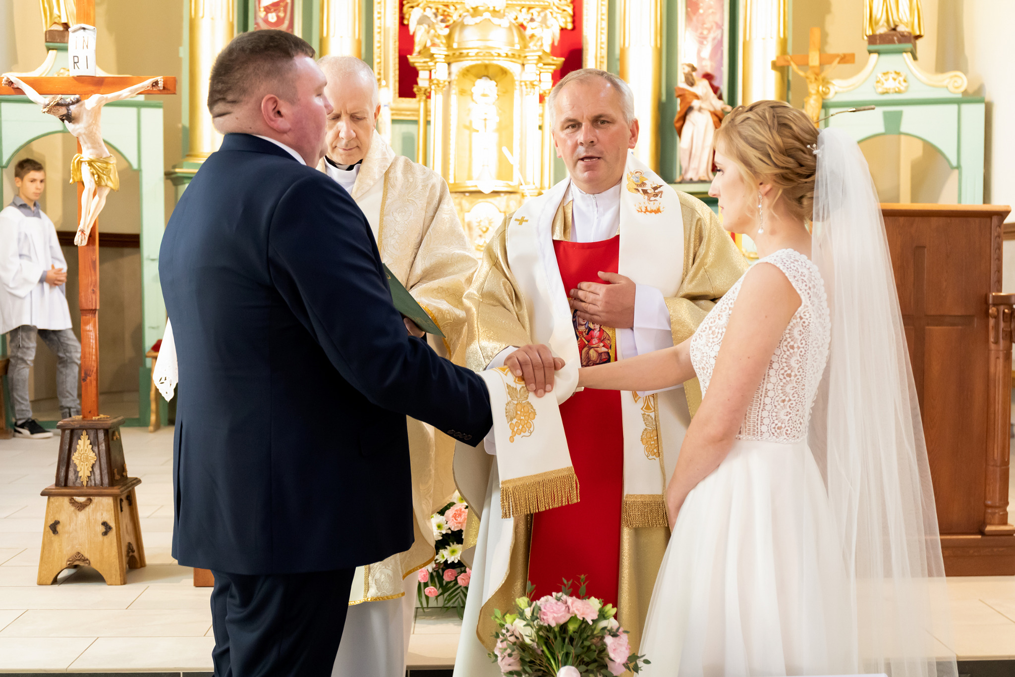 Uroczystość ślubna w kościele: para młoda wymienia obrączki w obecności księdza. W tle widoczny krucyfiks i ministrant. Jasne wnętrze kościoła.