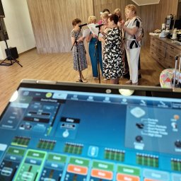 NEXA ENERGY Leszek Korycki - Grupa kobiet śpiewa do mikrofonów, widok zza konsolety mikserskiej. Jasne wnętrze z drewnianymi panelami. Sprzęt nagłośnieniowy w tle.