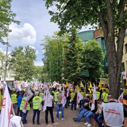 NEXA ENERGY Leszek Korycki - Zgromadzenie ludzi z flagami i transparentami na tle zieleni miejskiej i budynku. Uczestnicy w żółtych kamizelkach. Dzień słoneczny, widok na drzewa i budynki.