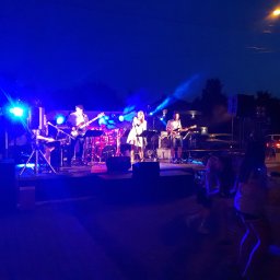 NEXA ENERGY Leszek Korycki - Zespół muzyczny na scenie plenerowej, oświetlony niebieskimi światłami, w tle publiczność. Wokalistka w białej sukience, gitarzyści i perkusista. Wieczorny koncert.