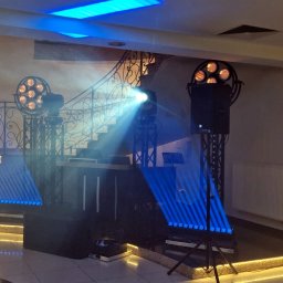 NEXA ENERGY Leszek Korycki - Scena z nagłośnieniem i oświetleniem na tle schodów z ozdobną balustradą. Widoczne reflektory, kolumny głośnikowe i konsoleta DJ-ska. Efekty świetlne w kolorze niebieskim.