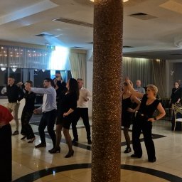 NEXA ENERGY Leszek Korycki - Grupa osób tańczy na parkiecie w eleganckiej sali balowej z kolumną. W tle widoczne stoły z gośćmi i dekoracje.