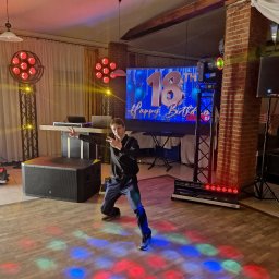 NEXA ENERGY DJ event Leszek Korycki - Mężczyzna w dynamicznej pozie na parkiecie, otoczony profesjonalnym sprzętem eventowym: nagłośnieniem, ruchomymi głowami, LED-ami i ekranem z napisem '18th Happy Birthday'.