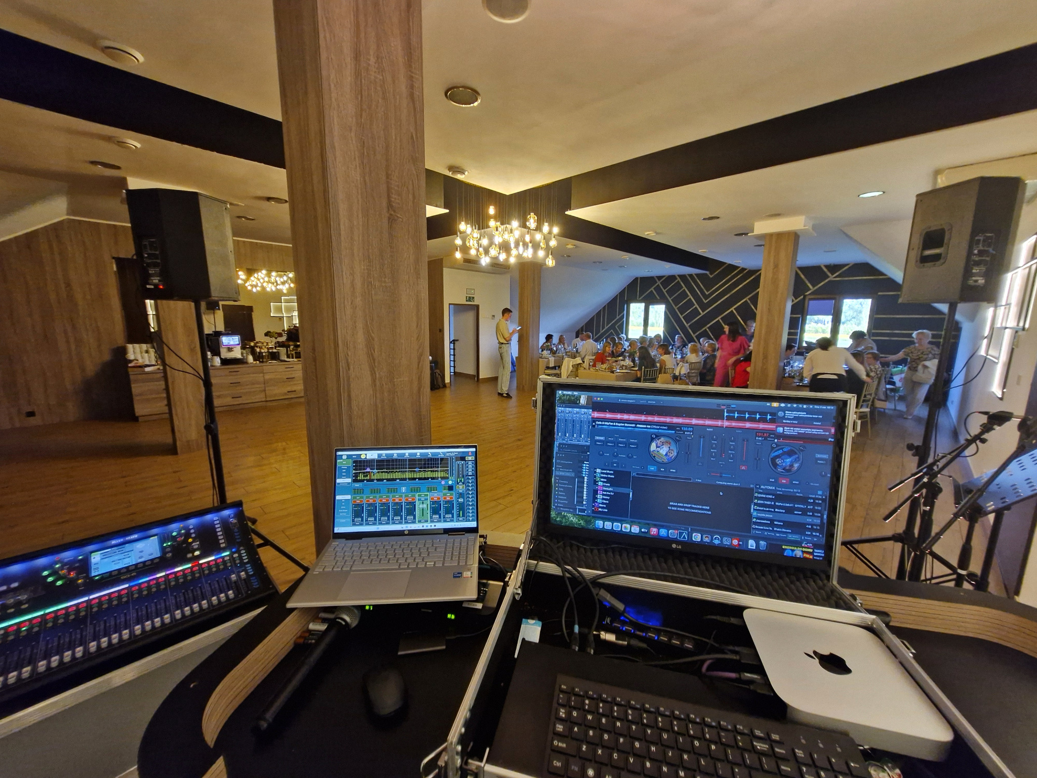 Sprzęt DJ-ski z laptopami i mikserem na tle sali bankietowej z gośćmi. Widoczne kolumny głośnikowe. Przygotowania do imprezy.