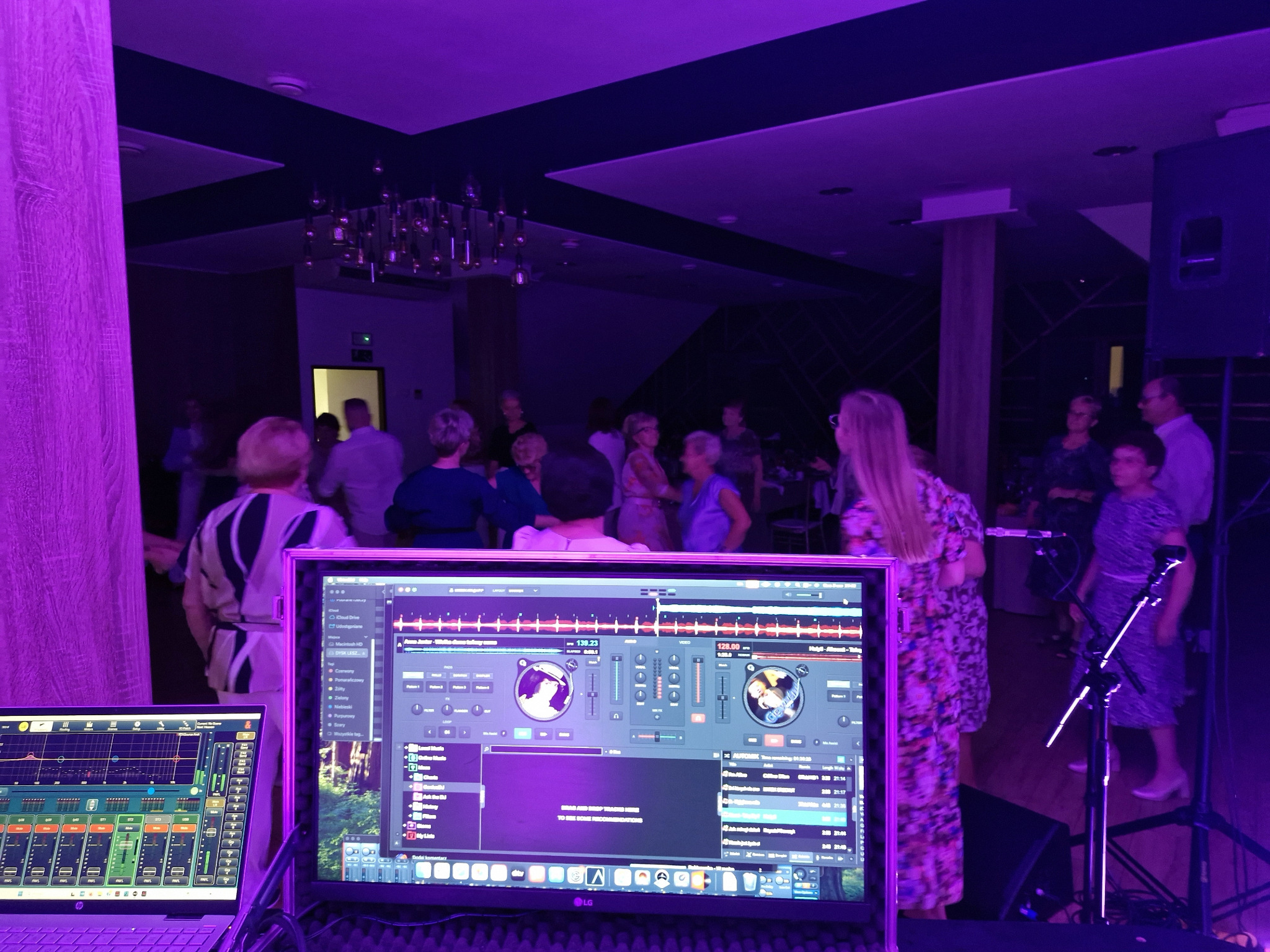 DJ grający muzykę na imprezie, widoczne monitory z oprogramowaniem DJ-skim, mikrofon i publiczność w tle, oświetlenie w odcieniach fioletu.