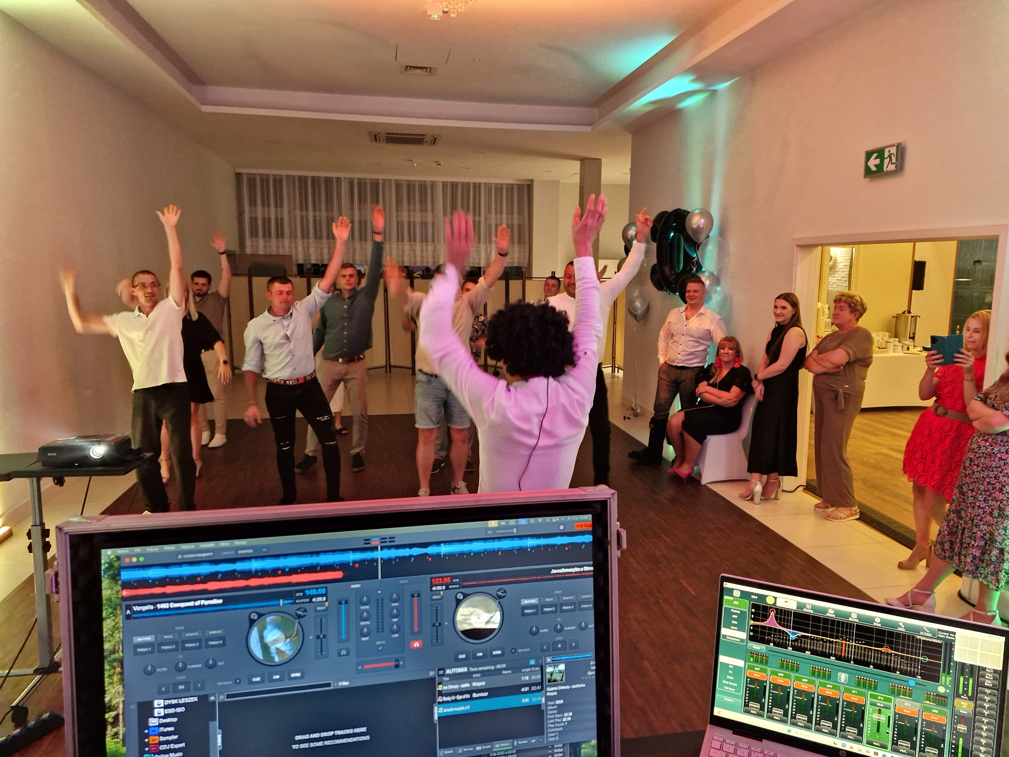 Parkiet i rozbawieni goście z podniesionymi rękami, DJ za konsoletą z widocznym oprogramowaniem. Dekoracje balonowe w tle. Impreza w jasnym, przestronnym wnętrzu.