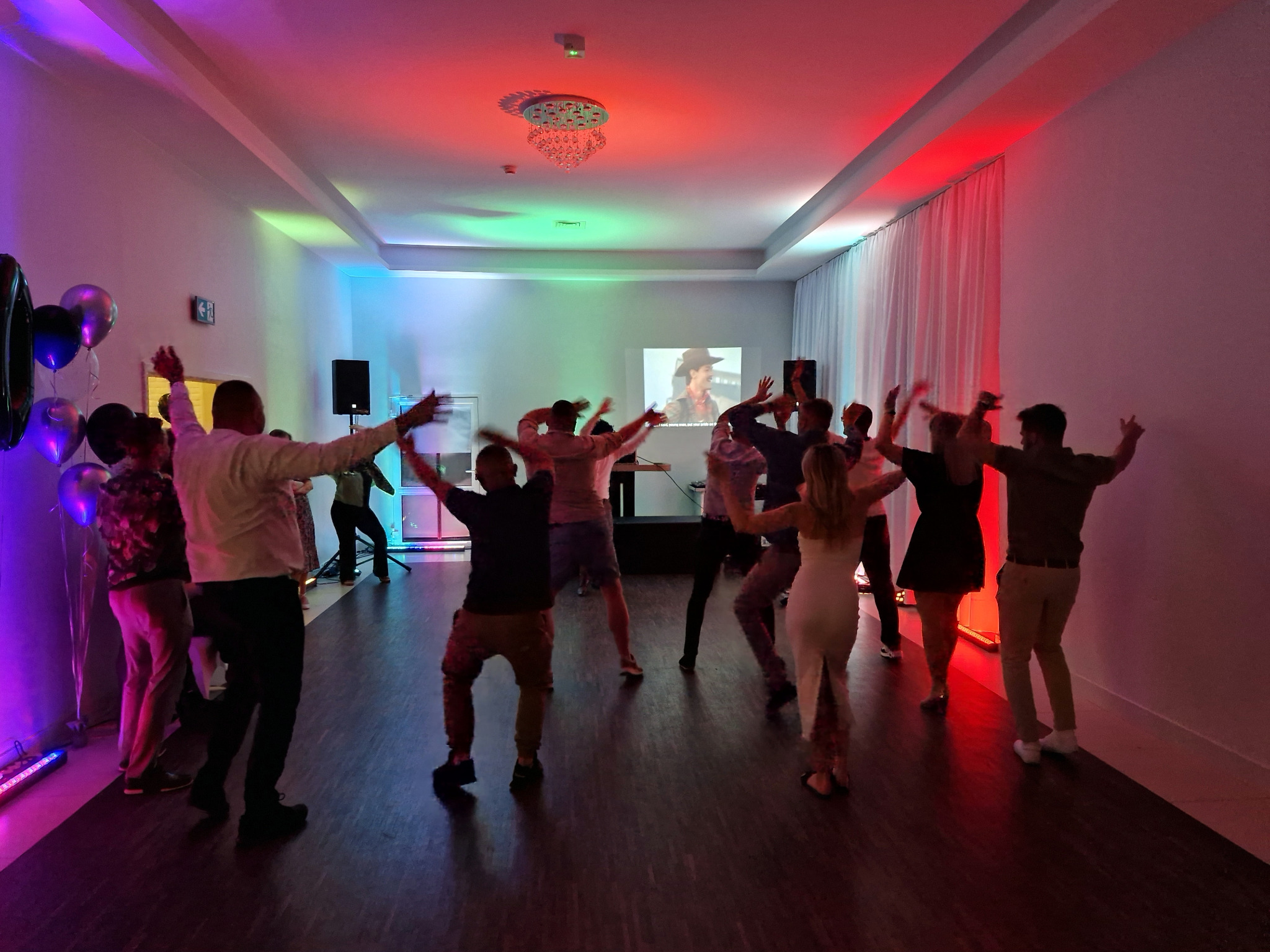 Roztańczona grupa osób w sali z kolorowym oświetleniem, balony i projektor w tle. Dynamiczna scena z imprezy integracyjnej.