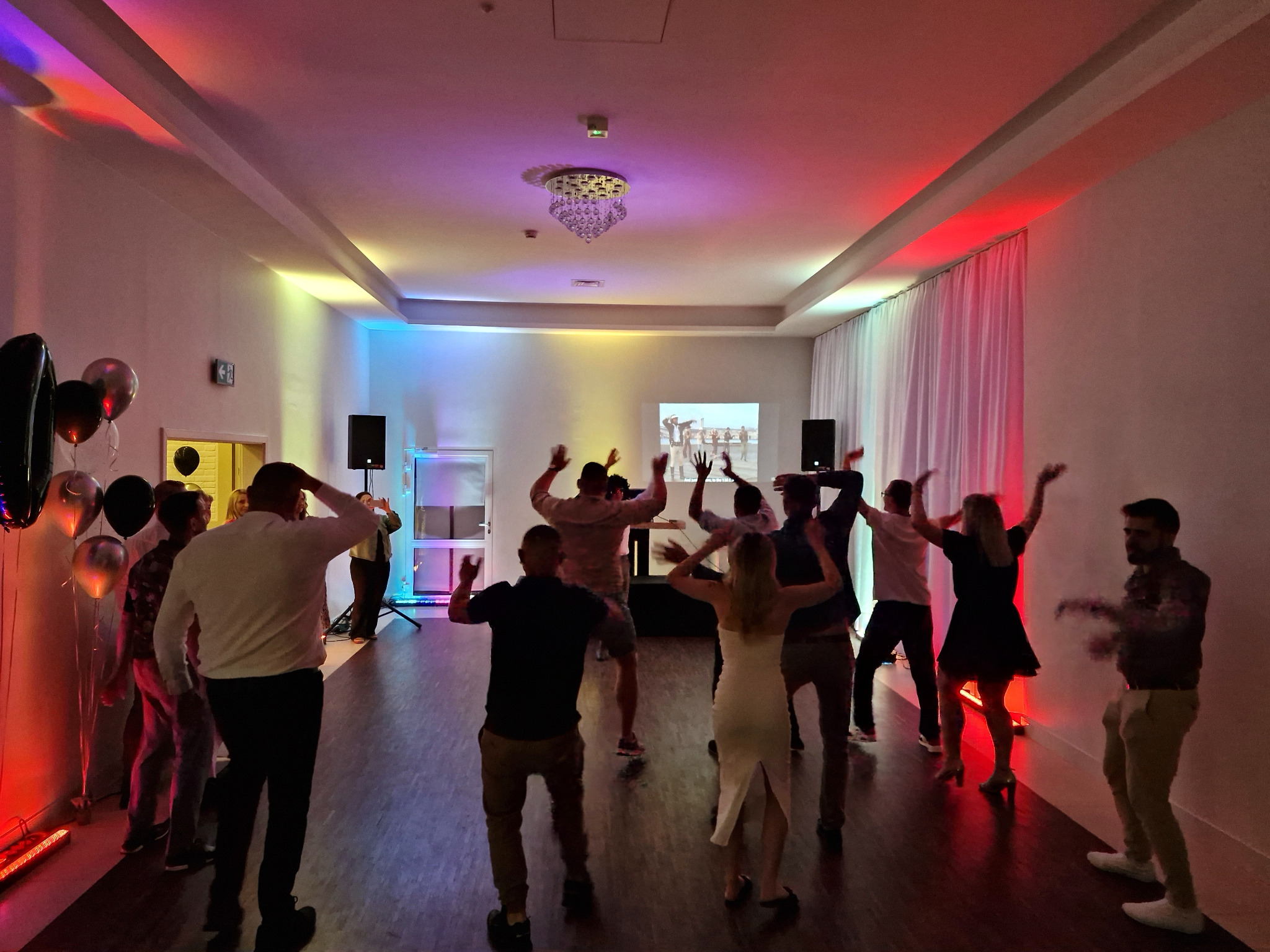 Rozbawieni goście tańczą w sali z kolorowym oświetleniem, widoczne balony i sprzęt nagłośnieniowy. Uczestnicy imprezy w ruchu, podnoszą ręce do góry.